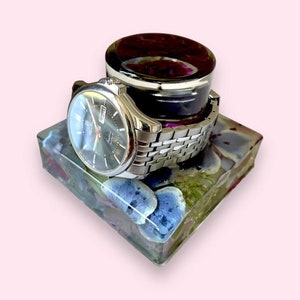 May include: A silver watch with a black face and a date window is resting on a square resin base with blue and purple flowers embedded inside. The watch is on top of a small black and purple resin lid.
