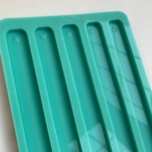 May include: A teal silicone mold with five rectangular cavities. Each cavity has a different design: a star, a heart, a teardrop, a crescent moon, and a leaf. The mold is labeled "Ata Molds".