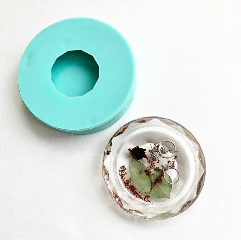 Puede incluir: Un plato para anillos de resina transparente con un borde facetado. El plato est&aacute; lleno de flores y hojas secas, y dos pendientes de plata descansan en la parte superior. El plato est&aacute; sobre una superficie blanca.