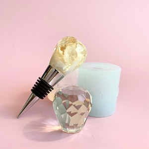 Puede incluir: Un tapón de botella plateado con una parte superior de resina transparente que contiene flores blancas secas y pan de oro. El tapón está al lado de un pisapapeles de cristal transparente en forma de diamante y un objeto cilíndrico azul claro.