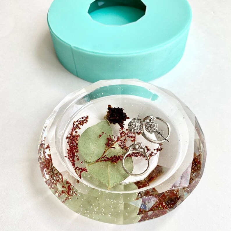 Puede incluir: Un plato para anillos de resina transparente con un borde facetado. El plato est&aacute; lleno de flores y hojas secas, y hay dos anillos y un par de pendientes dentro.