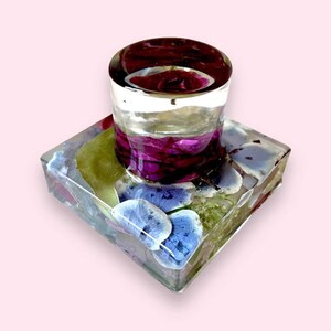 May include: A clear glass paperweight with a square base and a cylindrical top. The base contains pressed flowers in shades of blue, green, and purple. The top is filled with a clear liquid and has a layer of purple flowers.