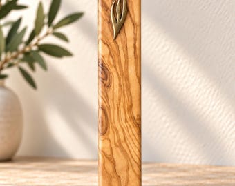 Puede incluir: Una caja de mezuzá rectangular hecha de madera de olivo pulida con un patrón de veta natural distintivo. La parte frontal presenta un pequeño emblema metálico en forma de llama cerca de la parte superior y dos orificios de montaje.