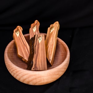 May include: A wooden bowl holds four wooden mezuzah cases. The cases are rectangular with a natural wood grain and a gold-colored emblem. The bowl and cases are made of light-colored wood, set against a black background.