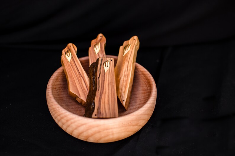 May include: A wooden bowl holds four wooden mezuzah cases. The cases are rectangular with a natural wood grain and a gold-colored emblem. The bowl and cases are made of light-colored wood, set against a black background.