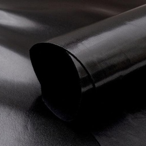 Könnte beinhalten: Ein großes, glattes, schwarzes Lederstück mit glänzender Oberfläche. Das Material ist oben aufgerollt und gibt den Blick auf eine dunkle, matte Unterseite frei. Ein weißes geometrisches Logo in Form eines stilisierten Würfels befindet sich unten rechts.