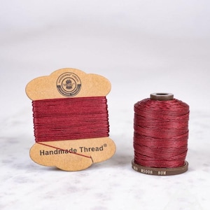 Può includere: Due rocchetti di filo color bordeaux. Uno è su un supporto di cartone con la scritta "Handmade Thread". L'altro è cilindrico con una base marrone. Il filo è ben avvolto su entrambi i rocchetti. Sul rocchetto cilindrico c'è la scritta "80M".