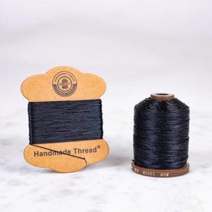Op de afbeelding: Twee spoelen zwart handgemaakt garen. Eén spoel zit op een bruine, wolkvormige kartonnen houder met de tekst "Handmade Thread". De andere is cilindrisch met een bruine basis en bovenkant. Ideaal voor naaien.