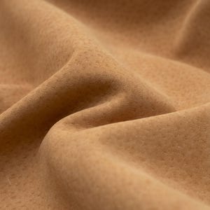 Può includere: Primo piano di un tessuto beige con una superficie strutturata e puntinata. Il tessuto è piegato, creando ombre e riflessi che enfatizzano la sua consistenza morbida ed elastica. L'aspetto generale suggerisce un materiale di alta qualità.