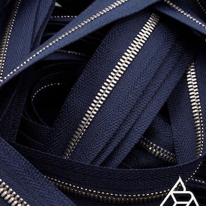 May include: Close-up of navy blue zippers with silver metal teeth. The zippers are arranged in a pile, showcasing the texture and detail of the fabric and metal components. The image highlights the contrast between the dark blue material and the shiny silver teeth.