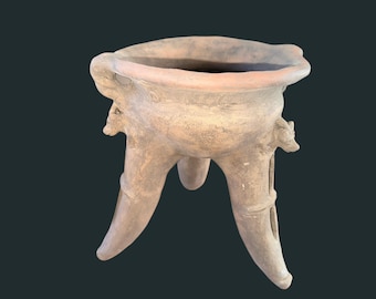 Puede incluir: Un recipiente de cerámica marrón claro con tres patas y un borde ancho e irregular. El recipiente tiene forma de cuenco redondeado y está adornado con pequeñas figuras con forma de animales en los laterales. Las patas son gruesas y curvas.