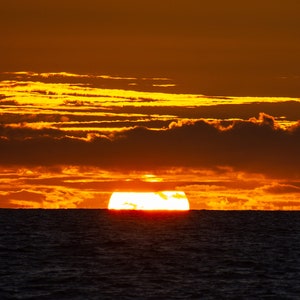 May include: A vibrant orange sunset over a dark body of water. The sun is setting behind the horizon, casting a warm glow on the clouds.