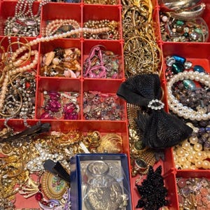 Pode incluir: Um organizador de joias vermelho dividido, cheio de v&aacute;rios colares, pulseiras, broches e brincos. As joias incluem pe&ccedil;as de ouro, prata, p&eacute;rolas e pedras preciosas. Uma fita preta com um centro de strass tamb&eacute;m &eacute; vis&iacute;vel.