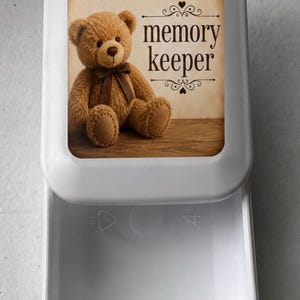 May include: A white plastic storage box with a sliding drawer. The lid features a printed image of a brown teddy bear with a dark brown ribbon around its neck, sitting on a wooden surface, with the text "memory keeper" in dark brown font.