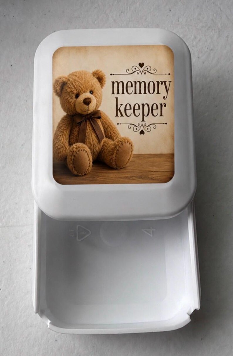 May include: A white plastic storage box with a sliding drawer. The lid features a printed image of a brown teddy bear with a dark brown ribbon around its neck, sitting on a wooden surface, with the text "memory keeper" in dark brown font.