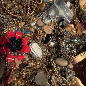 Pode incluir: Uma cole&ccedil;&atilde;o de joias vintage, incluindo colares, pulseiras, an&eacute;is e broches. As pe&ccedil;as s&atilde;o de v&aacute;rios estilos e cores, com tons dourados e prateados, e algumas com pedras preciosas e detalhes em esmalte. Uma broche de flor vermelha e preta &eacute; vis&iacute;vel.