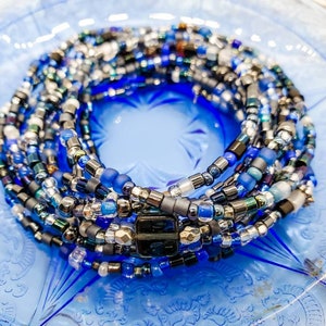 Puede incluir: Una colección de cuentas de cintura en tonos de azul, plata y negro. Las cuentas están dispuestas en un patrón en espiral, con una mezcla de acabados mate y brillantes. Las cuentas se exhiben en un plato de vidrio azul.