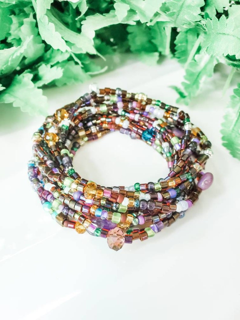Peut inclure: Bracelet multicolore en perles avec un charme en cristal. Le bracelet est compos&eacute; de petites perles rondes dans des tons de brun, violet, vert et or. Le charme en cristal est de couleur violet fonc&eacute;.