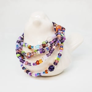 May include: Multicolored beaded bracelet with shades of purple, pink, orange, green, and blue. The bracelet is made of small glass beads and is wrapped around a white ceramic bird figurine.