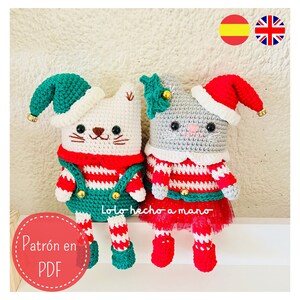 May include: Two crocheted cat elf toys, one white and one gray, wearing red and white striped outfits with green and red accents. The white cat elf is wearing a green elf hat and the gray cat elf is wearing a red Santa hat. The toys are sitting on a white shelf. The text "Patrón en PDF" is visible in the image.