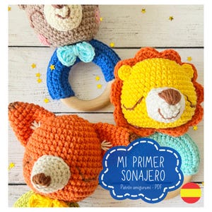 Op de afbeelding: Een set van drie gehaakte dierenrammelaars, waaronder een bruine beer, een gele leeuw en een oranje vos. De speelgoedjes zijn bevestigd aan een houten ring. De tekst "Mi Primer Sonajero" staat in blauw op een witte achtergrond met een gestippelde rand. De tekst "Patrón amigurumi - PDF" staat in zwart onder de titel. Een Spaanse vlag bevindt zich in de rechterbenedenhoek.