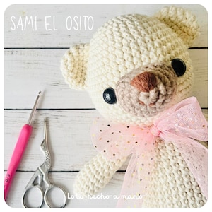 Op de afbeelding: Crèmekleurige gehaakte teddybeer met zwarte knoopogen en een bruine neus. Een roze strik met gouden glitteraccenten siert de nek van de beer. De tekst "SAMI EL OSITO" is zichtbaar. Een roze haaknaald en een zilveren schaar staan ook in de afbeelding.