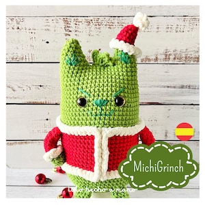 Puede incluir: Criatura verde de crochet parecida al Grinch que lleva un traje de Papá Noel rojo con ribetes blancos y un gorro de Papá Noel rojo y blanco. La criatura tiene ojos negros y una nariz verde cosida. El texto "MichiGrinch" está en un marco verde en forma de nube.