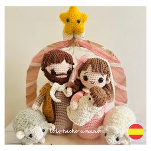 May include: A crocheted nativity scene with a yellow star, a brown-haired Joseph, a brown-haired Mary holding a baby Jesus, and two white sheep. The scene is set against a pink and brown background with the text "Lolo hecho a mano" in Spanish.