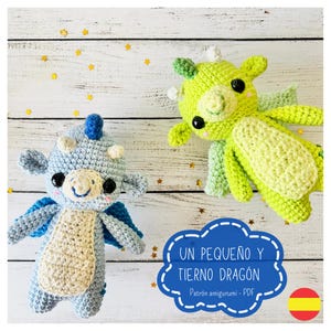 Op de afbeelding: Twee gehaakte amigurumi draken speelgoed, een blauwe en een groene, op een witte houten achtergrond. De tekst "UN PEQUEÑO Y TIERNO DRAGÓN Patrón amigurumi - PDF" is zichtbaar in de afbeelding.