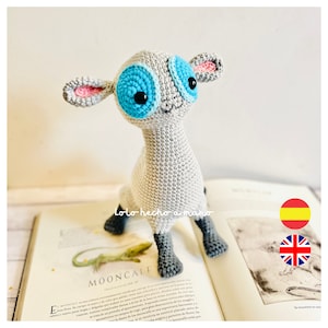 Op de afbeelding: Een grijs gehaakt amigurumi-speelgoed in de vorm van een lam met grote blauwe ogen en roze oren. Het speelgoed zit op een boek met de zichtbare tekst "MOONCALF". De tekst "Lolo hecho a mano" is ook zichtbaar op het speelgoed.