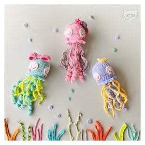 Könnte beinhalten: Drei gehäkelte Quallenspielzeuge in Pink, Blau und Grün mit gelben, pinken und grünen Tentakeln. Die Quallen befinden sich auf einem hellblauen Hintergrund mit kleinen weißen Punkten und gehäkelten Seetang.