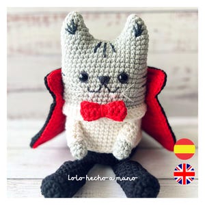 Puede incluir: Un gato de crochet gris vestido de vampiro con una capa roja y una pajarita roja. El gato está sentado sobre una superficie de madera blanca. El texto "Lolo hecho a mano" es visible en la pata del gato.
