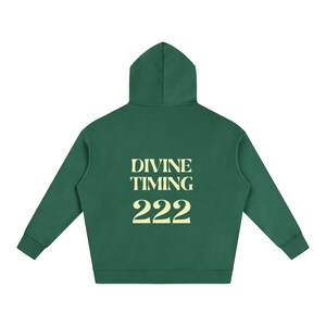 以下が含まれることがあります： 背面にライトクリーム色の文字で「DIVINE TIMING」と数字の「222」がプリントされた、フォレストグリーンのフード付きスウェットシャツ。長袖、フード、ストレートな裾が特徴で、平置きで中央に配置された状態で表示されています。