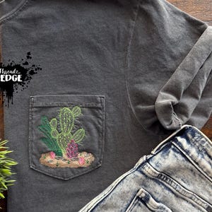 Puede incluir: Camiseta gris oscuro con un bolsillo bordado con un diseño de cactus en verde, rosa y marrón. La camiseta se combina con vaqueros de mezclilla con lavado ácido. El nombre de la marca "Threadz On Edge" es visible en el lado izquierdo de la camiseta.