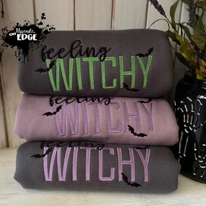Könnte beinhalten: Drei graue Sweatshirts übereinander gestapelt. Das oberste Sweatshirt hat den Text "feeling witchy" in Grün gestickt. Das mittlere Sweatshirt hat den Text "feeling witchy" in Lila gestickt. Das unterste Sweatshirt hat den Text "feeling witchy" in Lila gestickt. Jedes Sweatshirt hat eine schwarze Fledermaus auf jeder Seite des Textes gestickt.