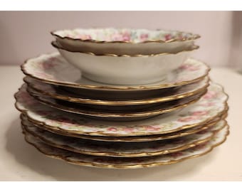 May include: A stack of six white porcelain plates with a pink and gold floral pattern. The plates have a scalloped edge and are decorated with gold trim.