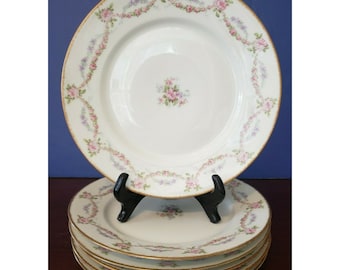 May include: A set of six white porcelain plates with a floral garland design in pink and purple, accented with gold trim. The plates are displayed on a black stand against a blue background, showcasing a classic, elegant aesthetic.