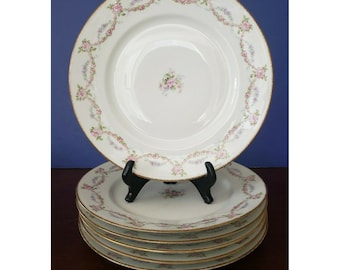 May include: Set of six white porcelain dinner plates with a delicate floral pattern in shades of pink and purple. The plates have a gold rim and are stacked on a wooden surface.