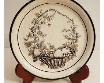 May include: A white ceramic dinner plate with a brown rim and a black and brown design of a basket of fruit and flowers.