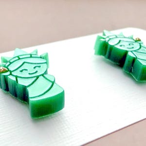 May include: A pair of stud earrings shaped like the Statue of Liberty in a translucent green material. Each earring features a smiling face, a crown with spikes, and a small gold-colored accent on the side, displayed on a white card.