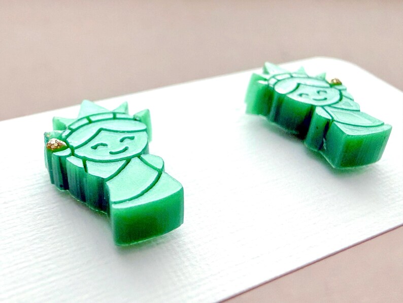 May include: A pair of stud earrings shaped like the Statue of Liberty in a translucent green material. Each earring features a smiling face, a crown with spikes, and a small gold-colored accent on the side, displayed on a white card.