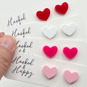 May include: Four pairs of heart-shaped earrings in red, white, pink, and light pink. The earrings are displayed on a white card with the text "Hooked & Happy".