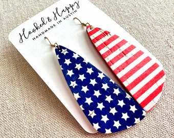 May include: A pair of cork earrings with a red, white, and blue American flag design. One earring features red and white stripes, while the other features blue with white stars. The earrings are displayed on a white card with the text "Hooked & Happy Handmade in Austin".