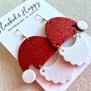 May include: Red and white glitter Santa hat earrings with gold hooks. The earrings are handmade in Austin, Texas. "Hooked & Happy" is written on the card.