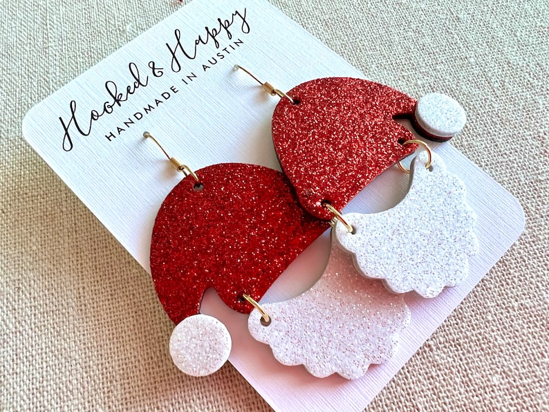 May include: Red and white glitter Santa hat earrings with gold hooks. The earrings are handmade in Austin, Texas. "Hooked & Happy" is written on the card.