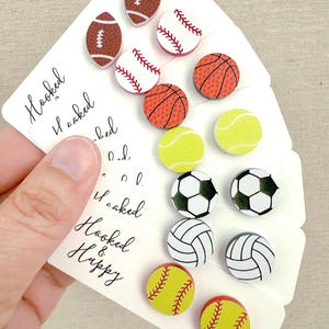 May include: A collection of sports-themed buttons featuring footballs, baseballs, basketballs, tennis balls, soccer balls, volleyballs, and softballs. The buttons are in various colors and designs, with the text "Hooked & Happy" on the card.