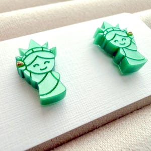 May include: A pair of stud earrings shaped like the Statue of Liberty, crafted from a smooth, light green material. Each earring features a stylized, smiling face with a crown and a small, gold-colored accent detail on the torch side.