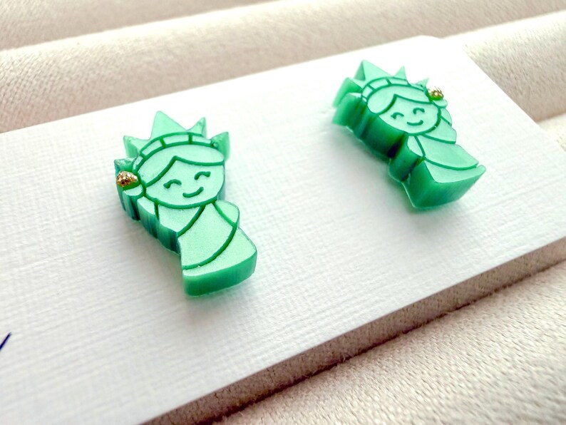 May include: A pair of stud earrings shaped like the Statue of Liberty, crafted from a smooth, light green material. Each earring features a stylized, smiling face with a crown and a small, gold-colored accent detail on the torch side.