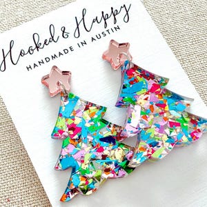 May include: A pair of festive Christmas tree earrings. The earrings are shaped like trees and filled with colorful confetti. Each earring has a pink star at the top. The earrings are on a white card that says "Hooked & Happy Handmade in Austin."