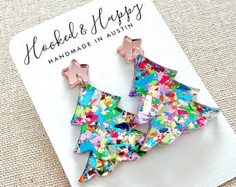 May include: A pair of festive Christmas tree earrings. The earrings are shaped like trees and filled with colorful confetti. Each earring has a pink star at the top. The earrings are on a white card that says "Hooked & Happy Handmade in Austin."